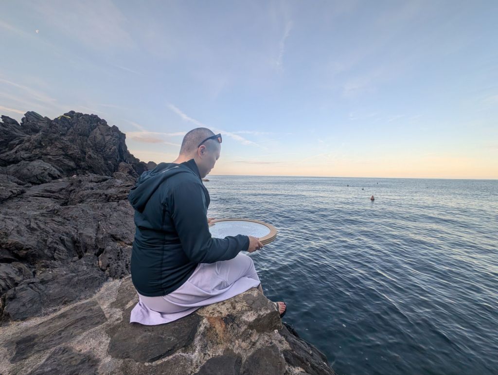 Picture of woman in dress with ocean drum, sitting by the ocean.